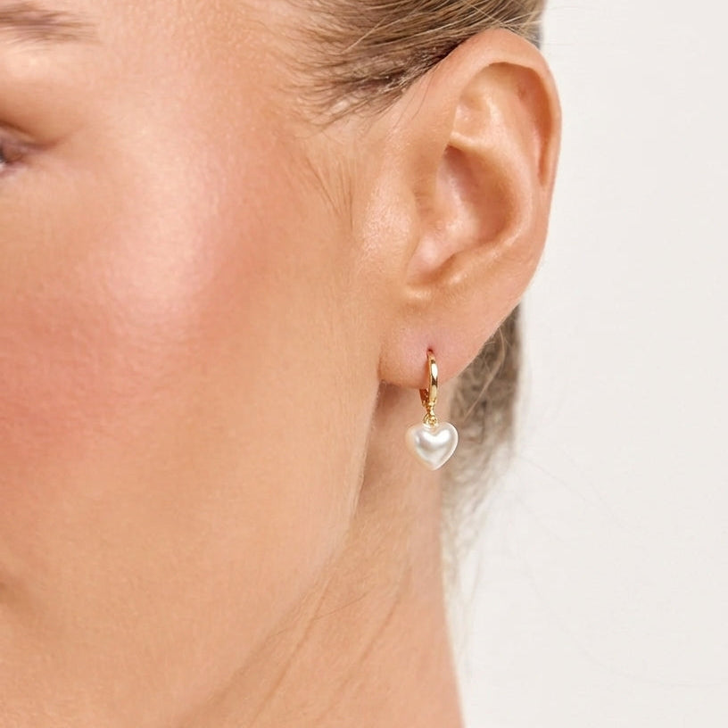 A close-up of a model's ear wearing a small gold huggie hoop earring with a dangling white heart-shaped pearl.