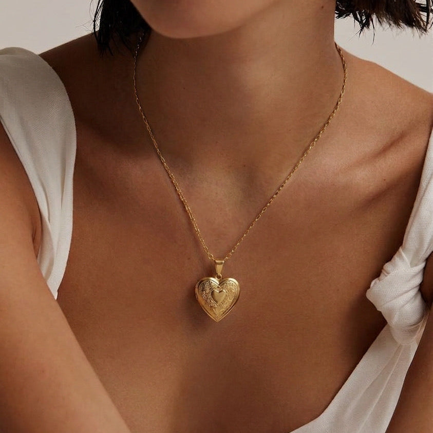 A close-up of a model wearing a delicate gold chain necklace with a textured heart-shaped pendant, styled with a white top against a neutral background.
