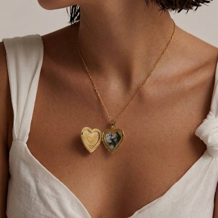 A close-up of a model wearing a delicate gold necklace with two heart-shaped pendants, one plain gold and the other featuring a small photo insert, styled with a white tied-strap top.